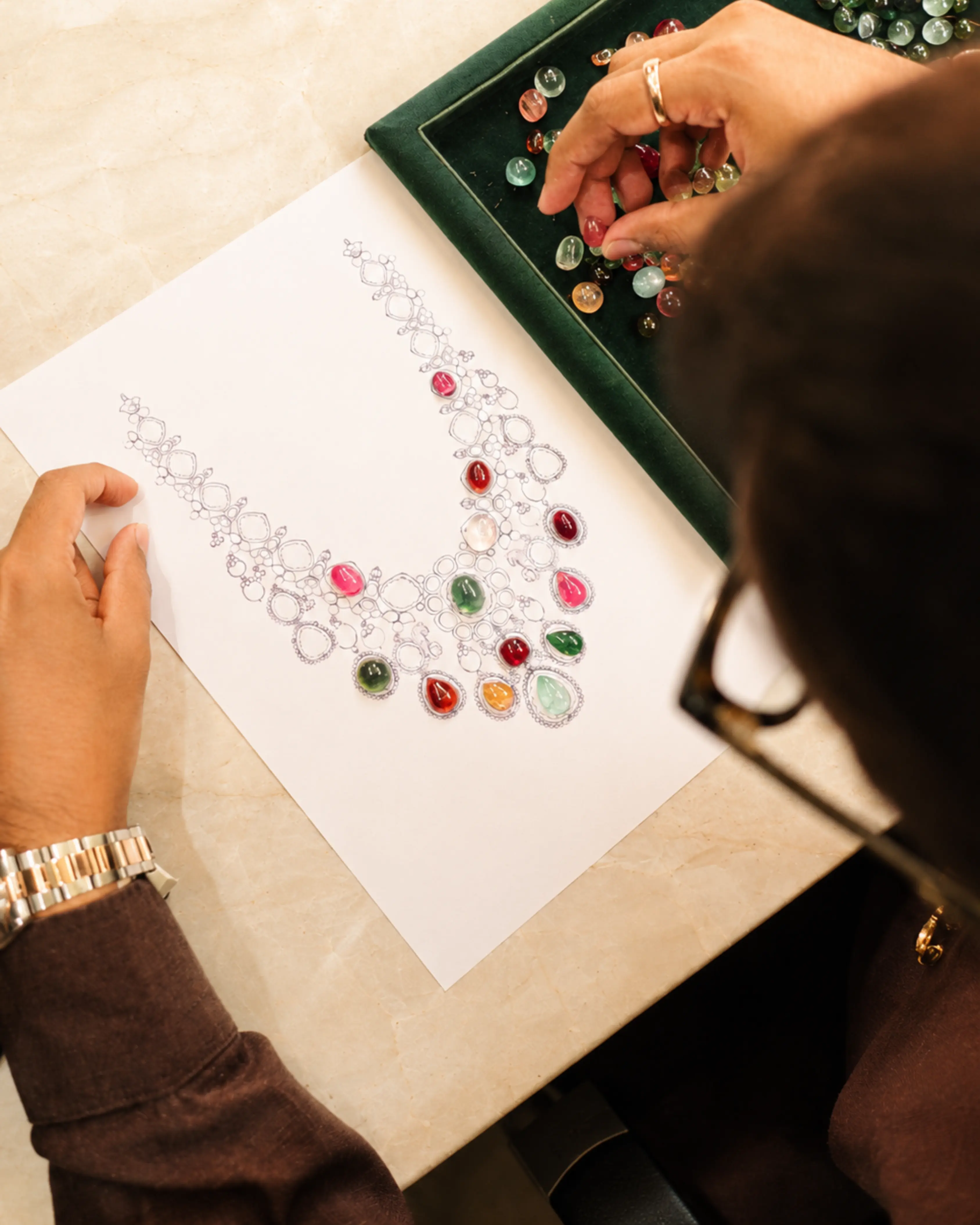 A Padmini designer choosing coloured gemstones beside a hand-drawn necklace sketch